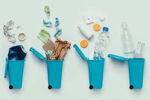 A flat lay of four blue trash cans laying on a white background with recycled items